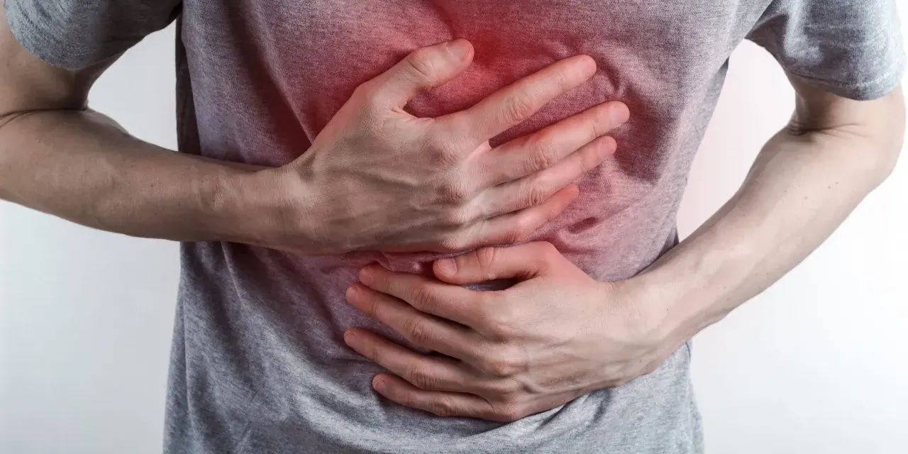 Osoba trzyma się za brzuch, odczuwając ostry ból w klatce piersiowej na środku. Czerwony obszar na koszulce sugeruje miejsce dolegliwości.