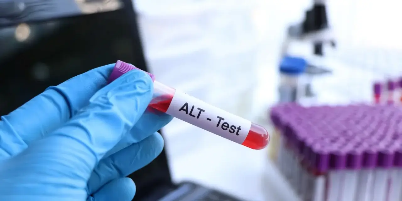 Ręka w niebieskiej rękawiczce trzyma probówkę z krwią oznaczoną "ALT - Test". W tle widać mikroskop i stojak z probówkami. To badanie ALT.