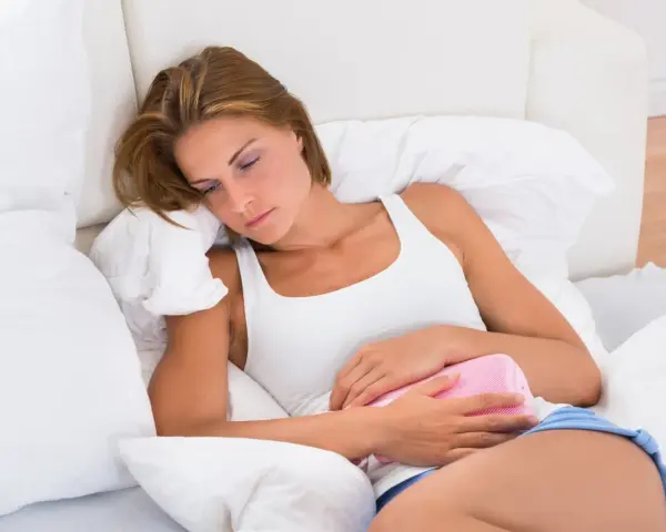 Kobieta z termoforem na brzuchu, cierpiąca z powodu bólu. To może być objaw PMS, co to właściwie oznacza?