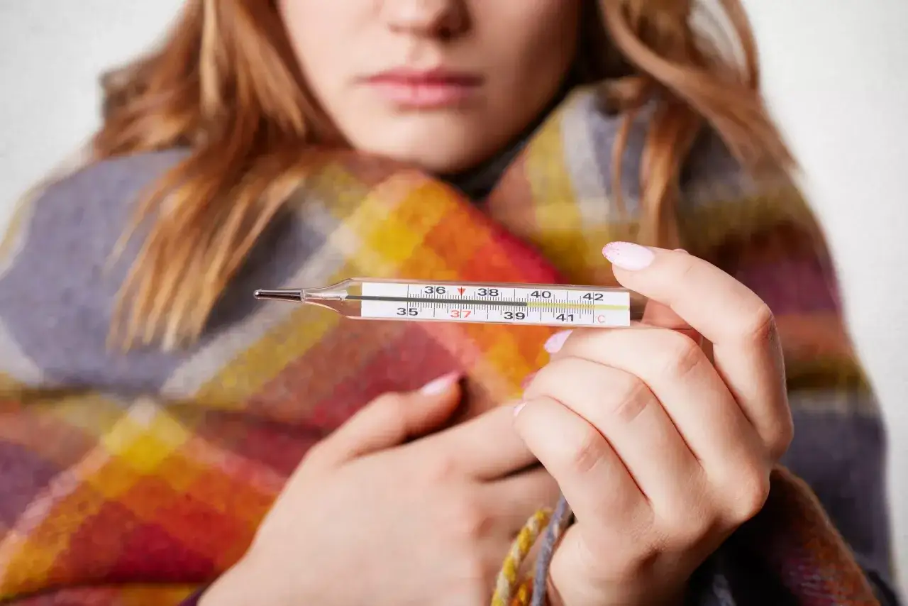 Kobieta z gorączką owinięta w koc trzyma termometr. Szuka informacji, jaki lek na covid może pomóc.